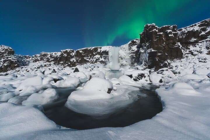 Oxararfoss Waterfall appears frozen as northern lights glow above it at night.